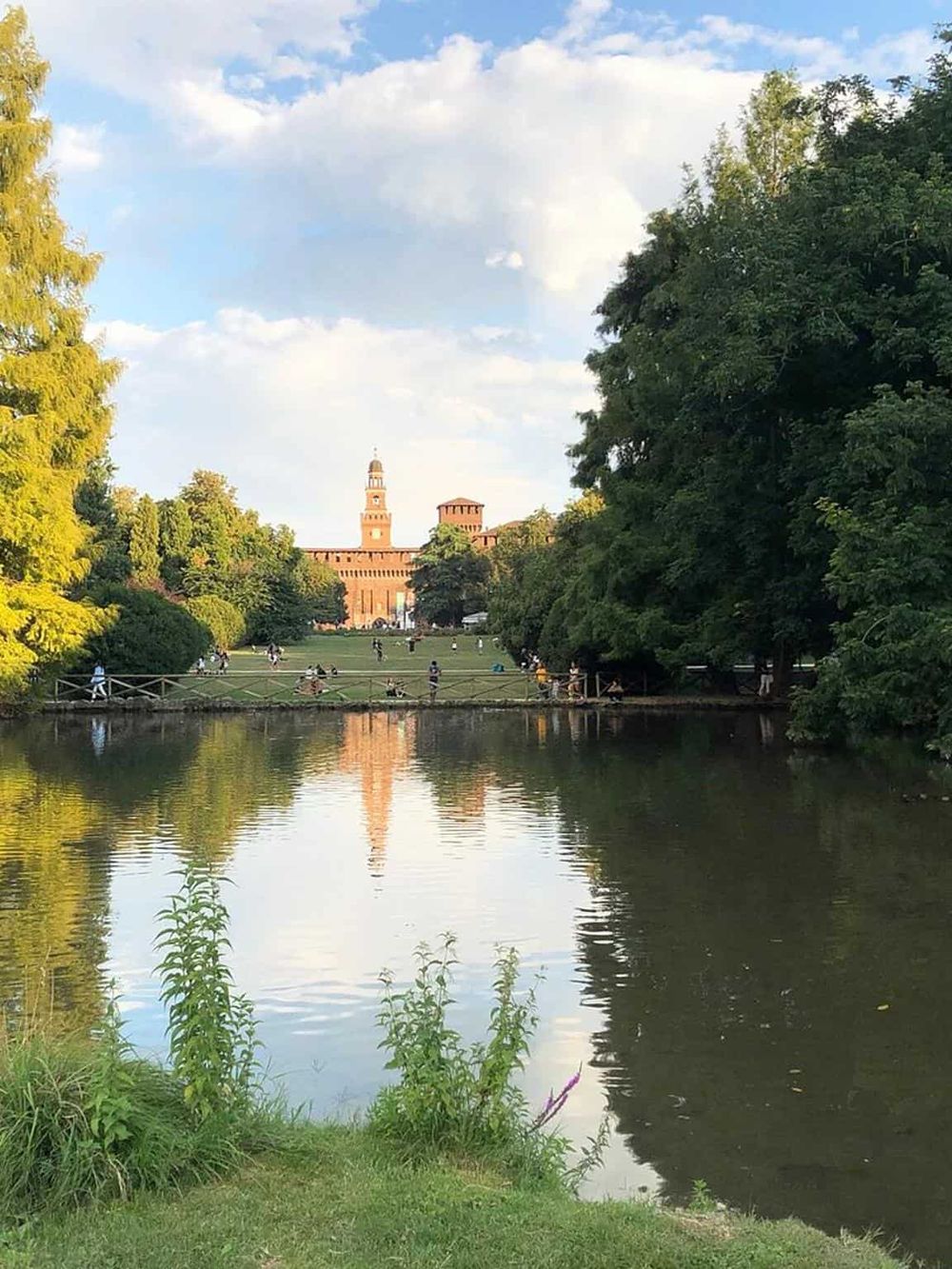 Serene park view with a historic brick castle and lush green trees reflected in calm water, perfect for relaxation and outdoor activities.