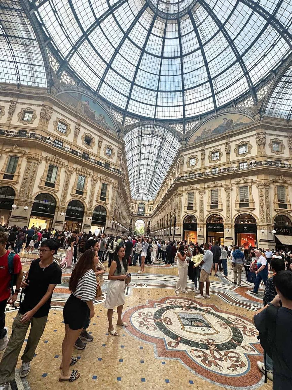 Vast shopping mall interior with ornate architecture, glass ceiling, and shoppers browsing luxury brands.
