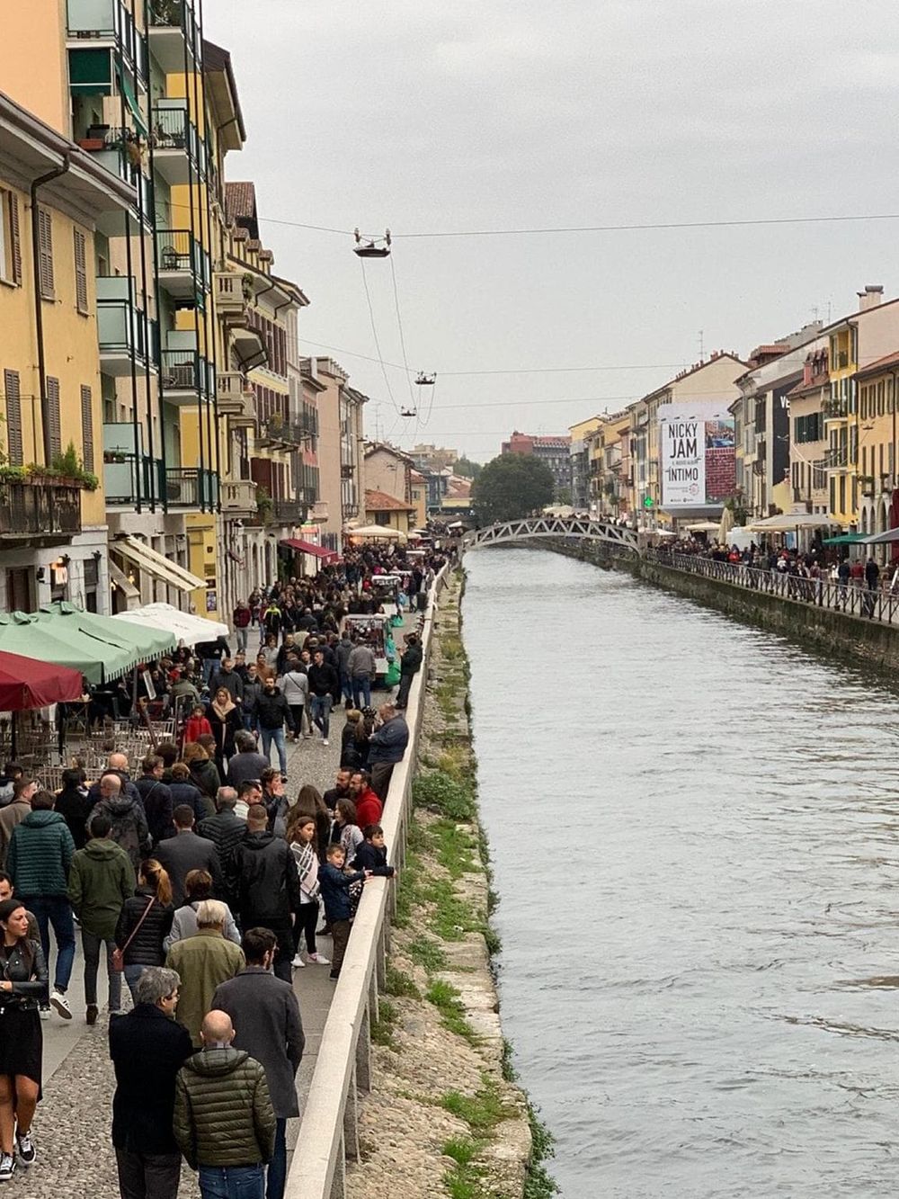 Bustling canal-side promenade in Italy with crowded outdoor cafes, colorful buildings, and scenic waterway.