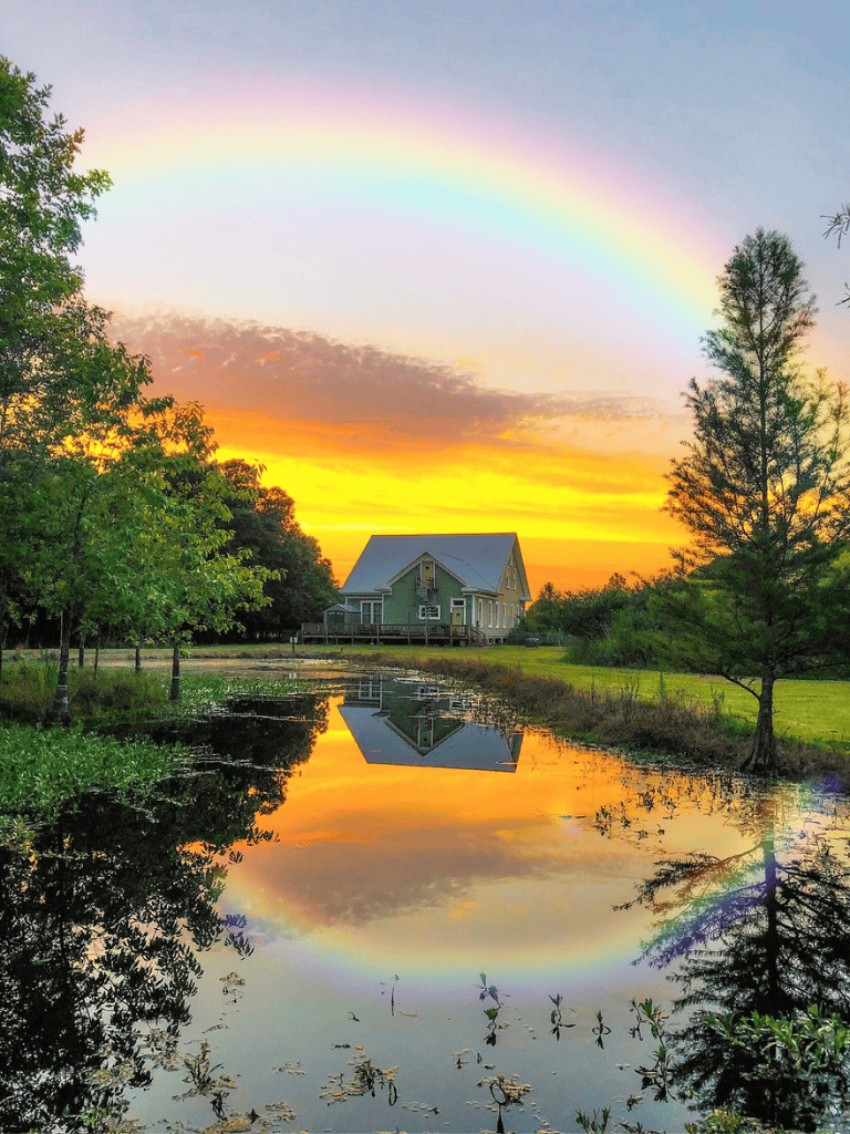 Colorful rainbow over serene pond with house, vibrant sunset, lush trees, and reflection in water, beautiful outdoor scenery.