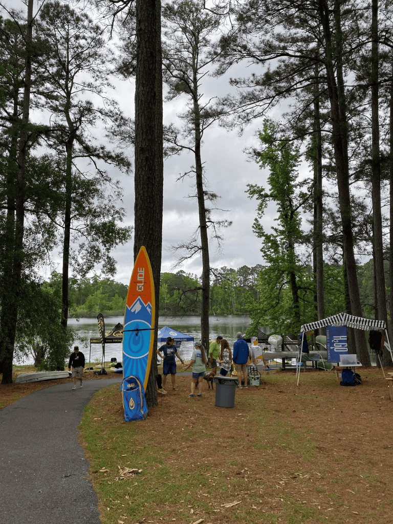 Surfboard and outdoor event setup at Quest for Directions event by a lake with trees and cloudy sky.