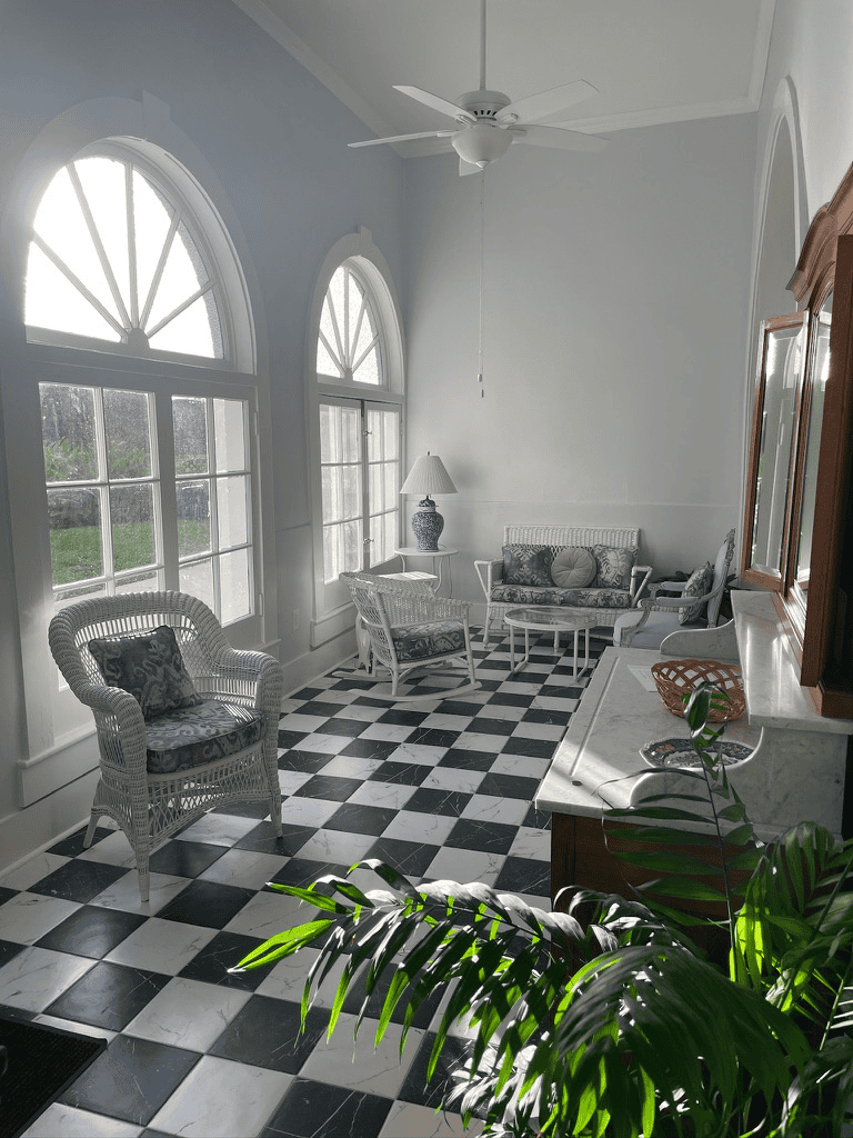 Bright sunlit sitting area with large arched windows and black-and-white checkered flooring, elegant wicker and upholstered furniture.