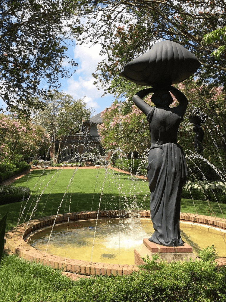 Elegant garden fountain with female statue carrying a large shell, surrounded by lush greenery and blooming trees.