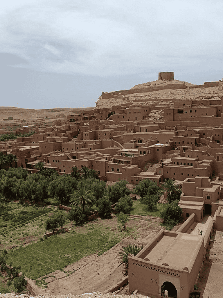 Ancient mud-brick village in the Moroccan desert, perched on a hillside with lush greenery in the foreground.