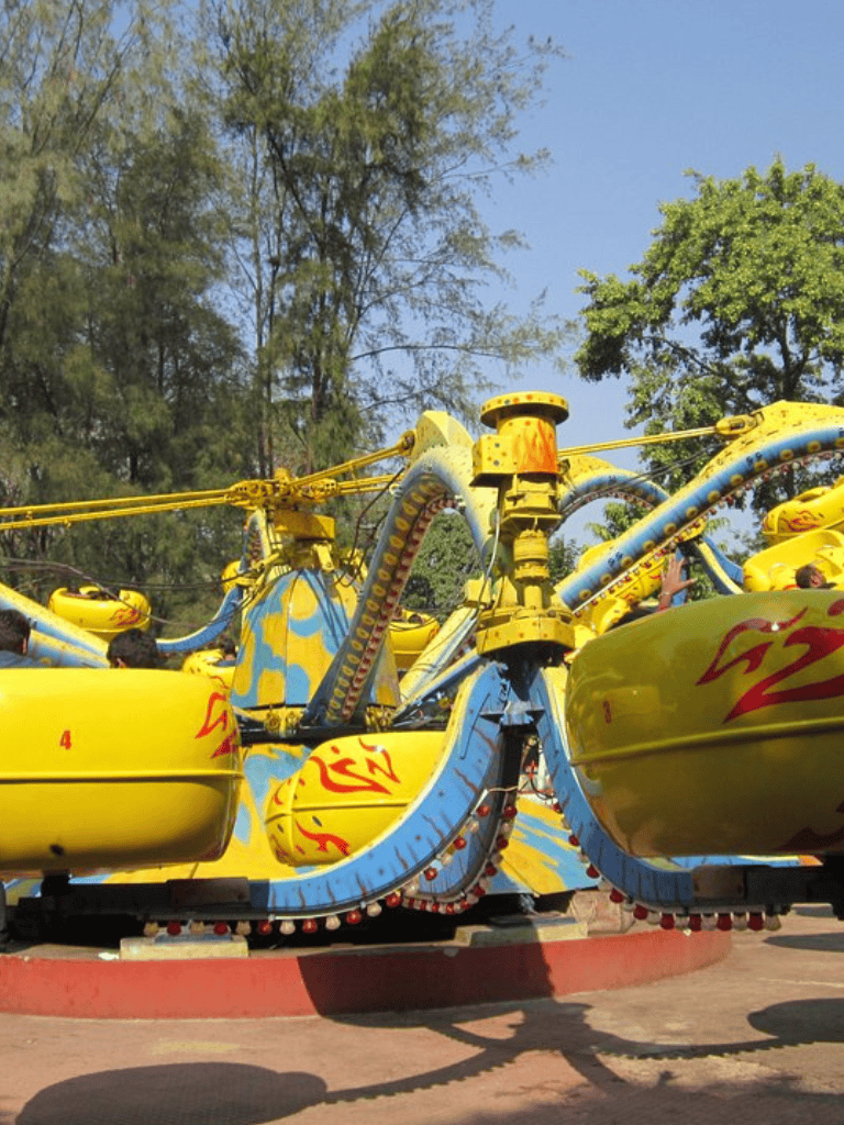 Children's amusement park ride with colorful yellow and blue spinning cars, outdoor setting with trees and clear sky.