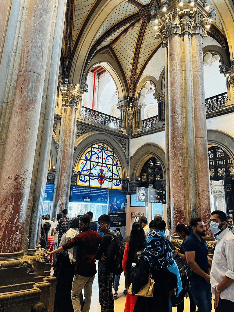 Grand Railway Station interior with travelers, ornate columns, stained glass, and vibrant architecture.