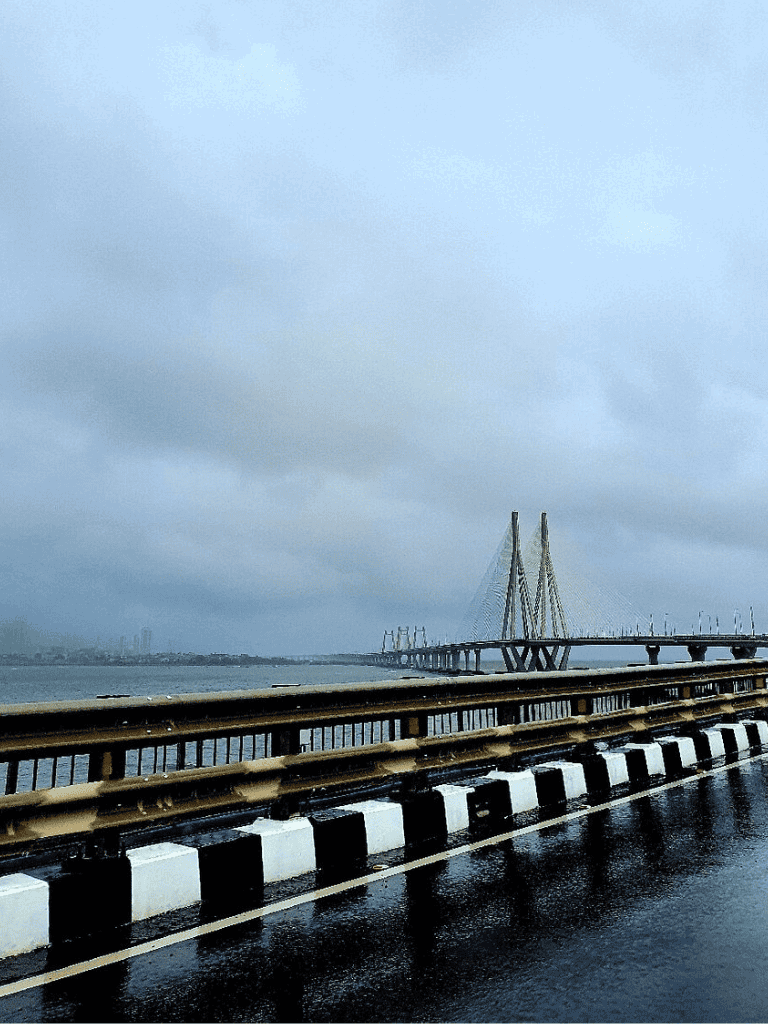 Overcast sky over a bridge and waterway, rainy road with black and white lane markers, city skyline in the background.