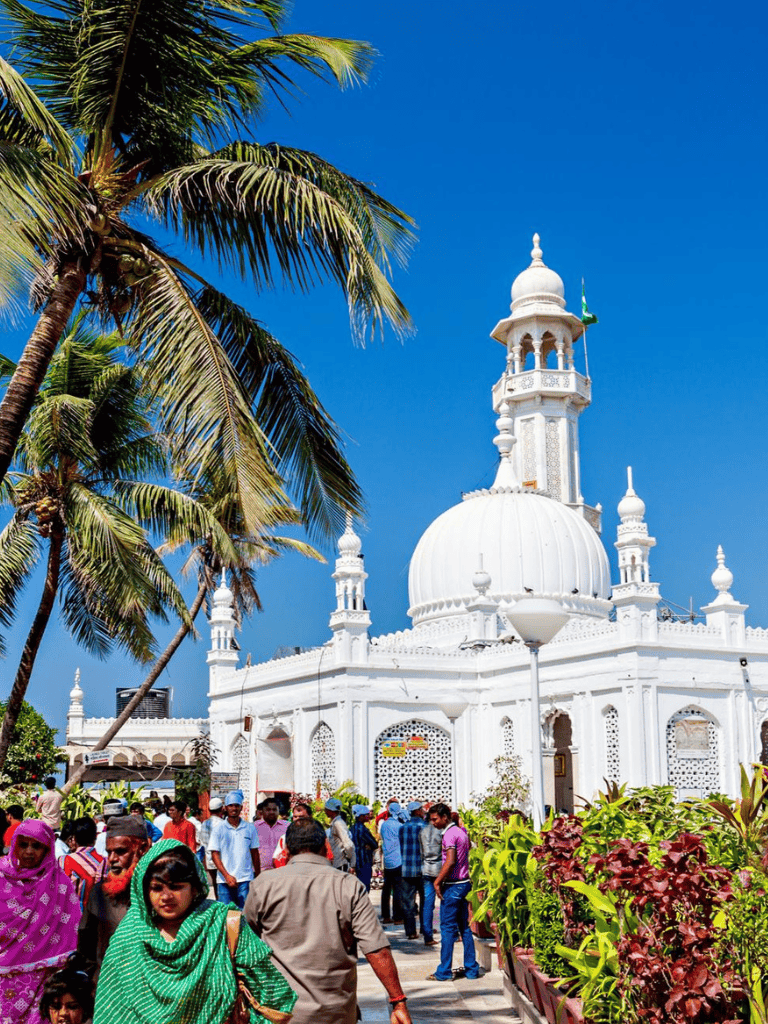 Colorful people walking outside white mosque with dome and minaret, under palm trees, sunny day in India.