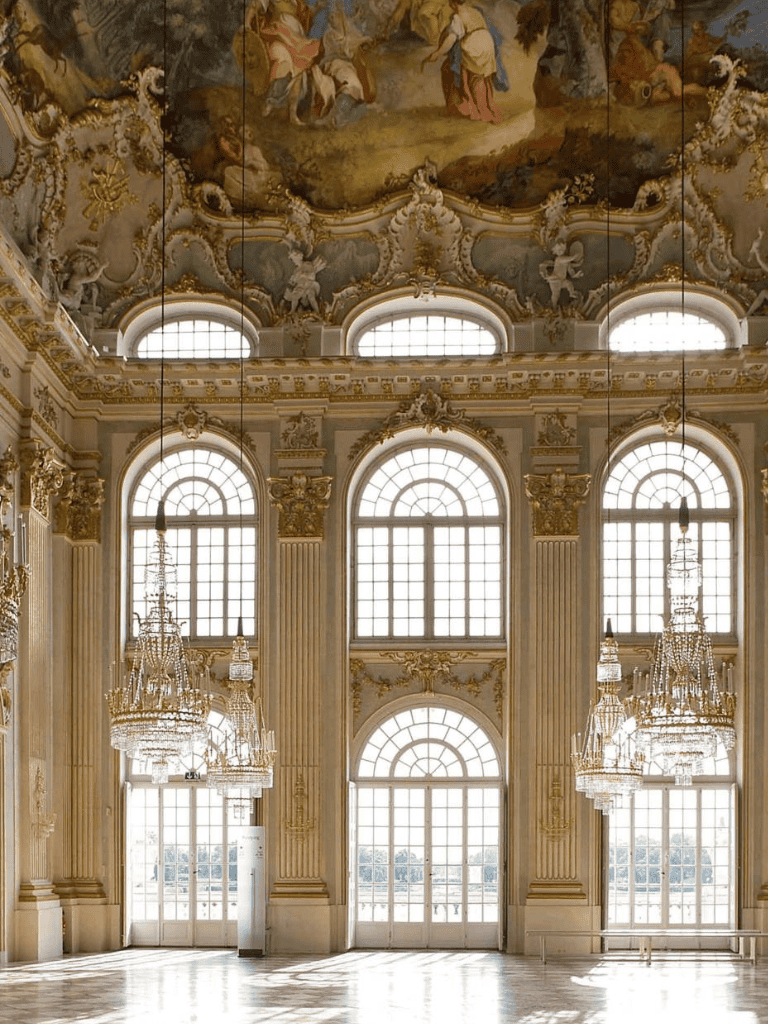 Opulent grand hall with large arched windows, ornate gold accents, and crystal chandeliers in a historic palace inside.