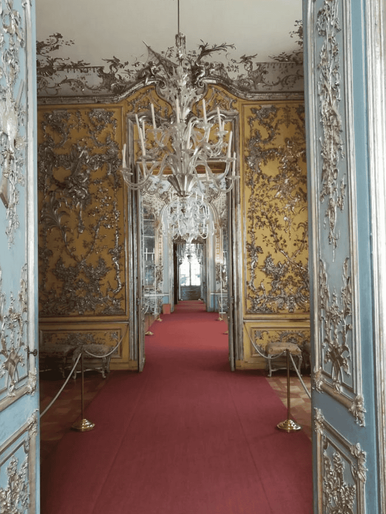 Elegant historic palace hallway with ornate gold and blue decor, grand chandelier, and luxurious red carpet.