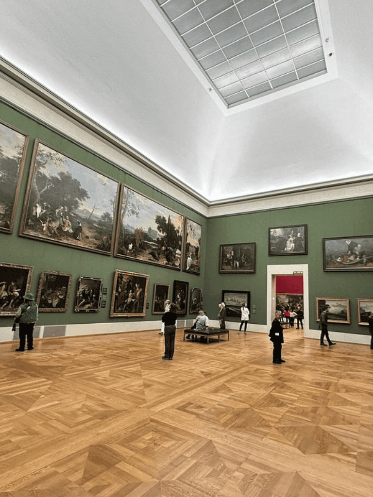 Alt text: Art gallery interior with framed paintings, wooden floor, and visitors viewing artwork.