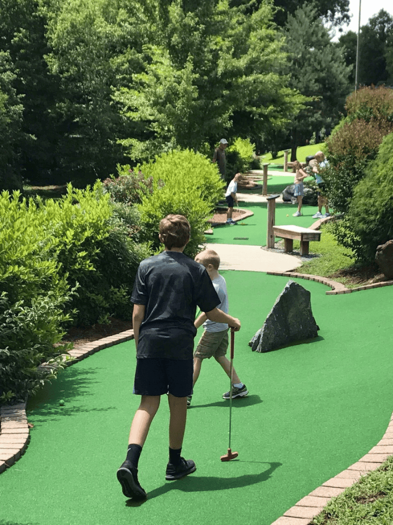 Mini golf game at lush outdoor course with children and adults enjoying fun family activity.