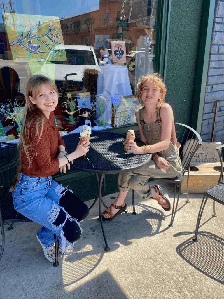 Sweet girls enjoying ice cream outside a cozy café with colorful storefront decor.