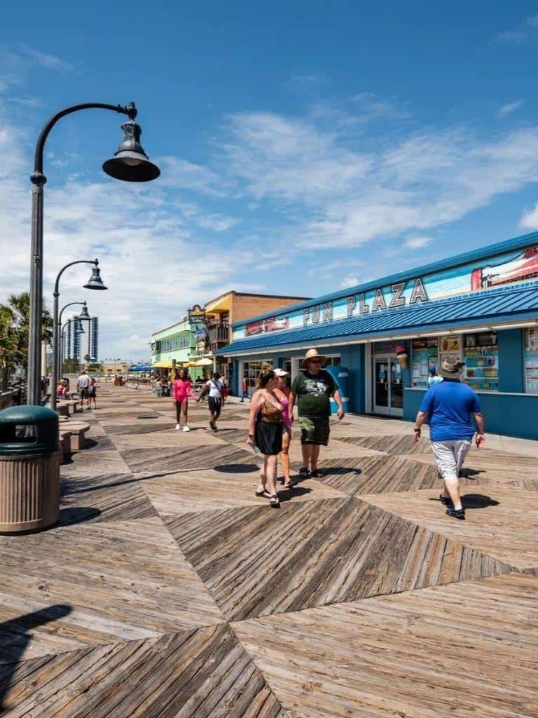 Brightly lit amusement park boardwalk with visitors, colorful shops, and a clear blue sky.