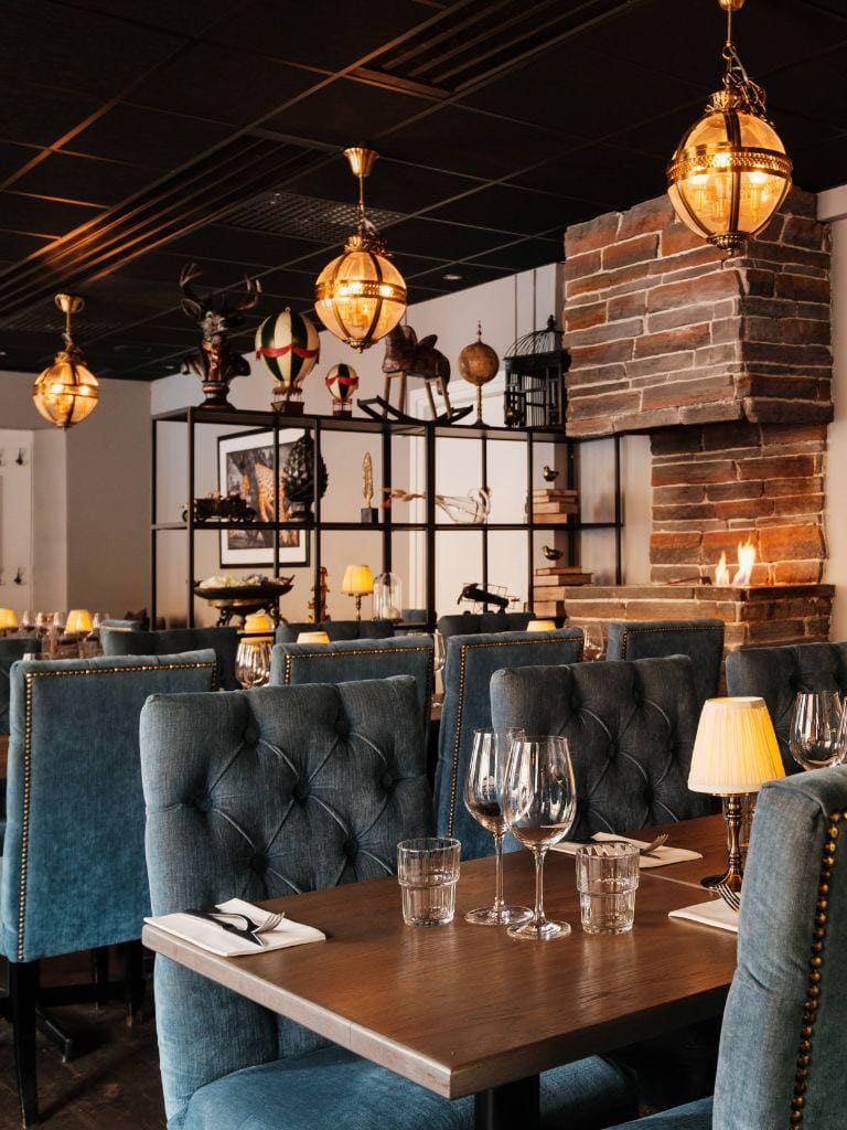 Elegant restaurant interior with brick fireplace, plush chairs, pendant lighting, and decorative shelves.