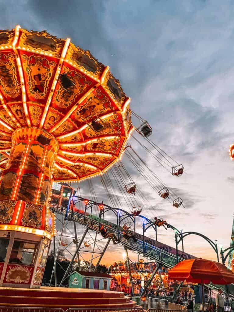 Amusement park swing ride at dusk with vibrant carnival lights and roller coaster in the background.