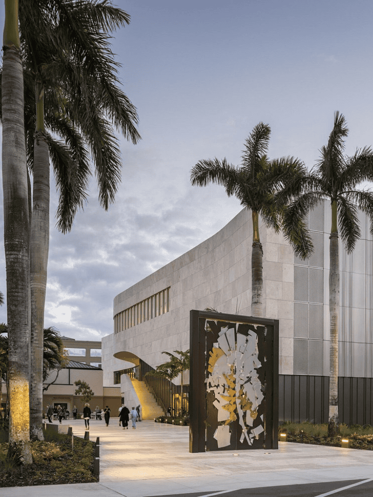 Modern architectural building with palm trees, outdoor seating, and artistic sculpture in a landscaped area.
