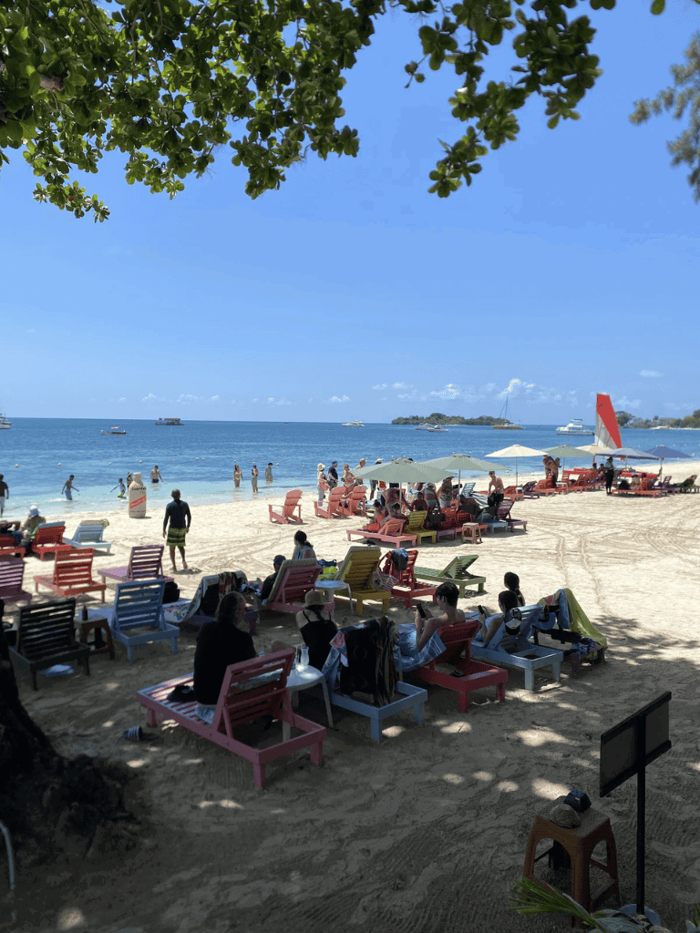 Sunlit beach scene with colorful lounge chairs, umbrellas, and visitors enjoying seaside relaxation.