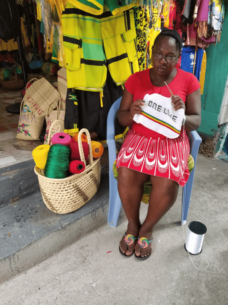 Stranded woman knitting colorful hat at market, vibrant clothing and craft supplies in background.