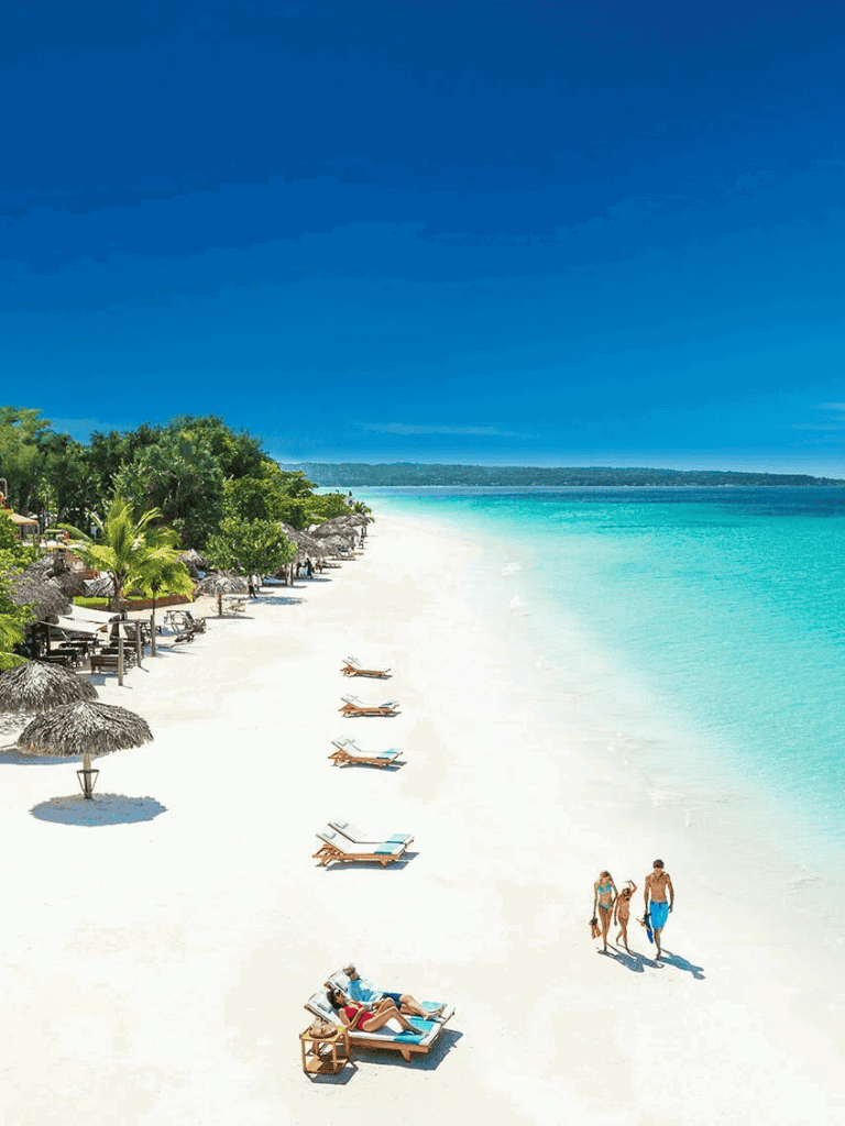 Tropical beach with white sand, clear turquoise water, and lounge chairs under straw umbrellas.