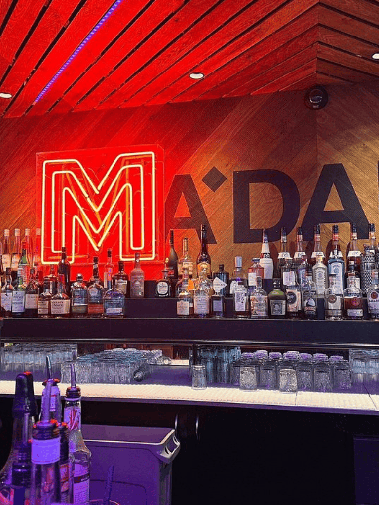 Neon M sign at stylish bar with liquor bottles and glassware, modern nightlife setting.