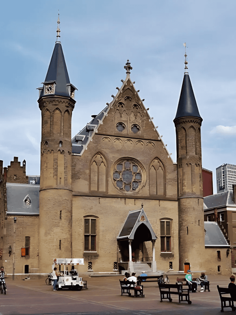 Old European castle with towers and Gothic architecture, located in an urban area with benches and pedestrians.