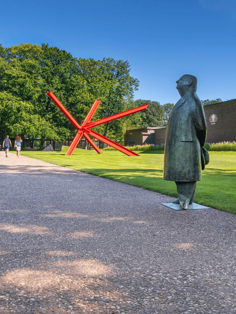 Bold orange modern outdoor clock sculpture with a metallic statue of a man looking up, set in a lush green park.