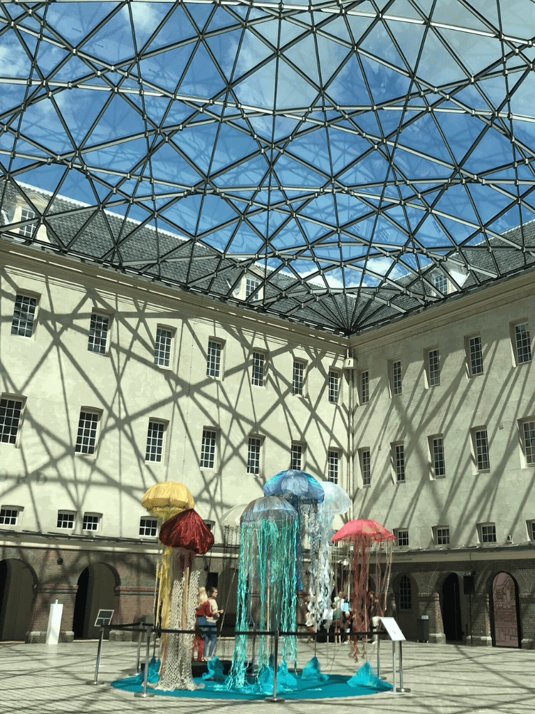 Colorful jellyfish art installation under a glass roof in a courtyard with shadows of geometric patterns.