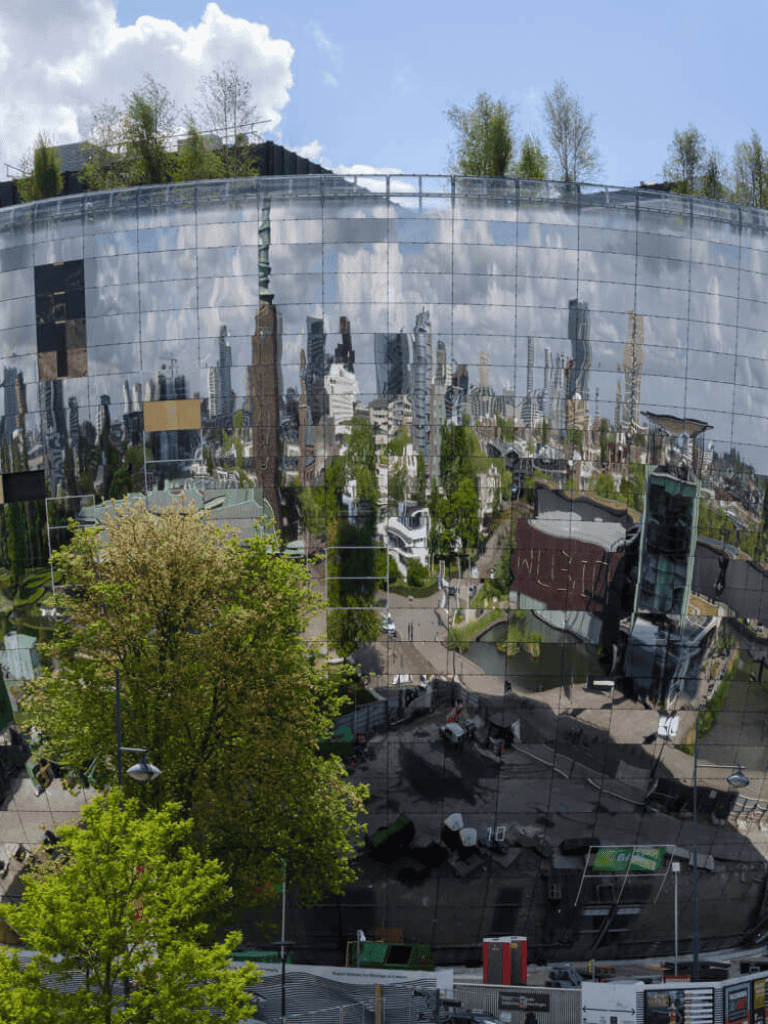 Shiny reflective building with cityscape mirror image and green trees in foreground.