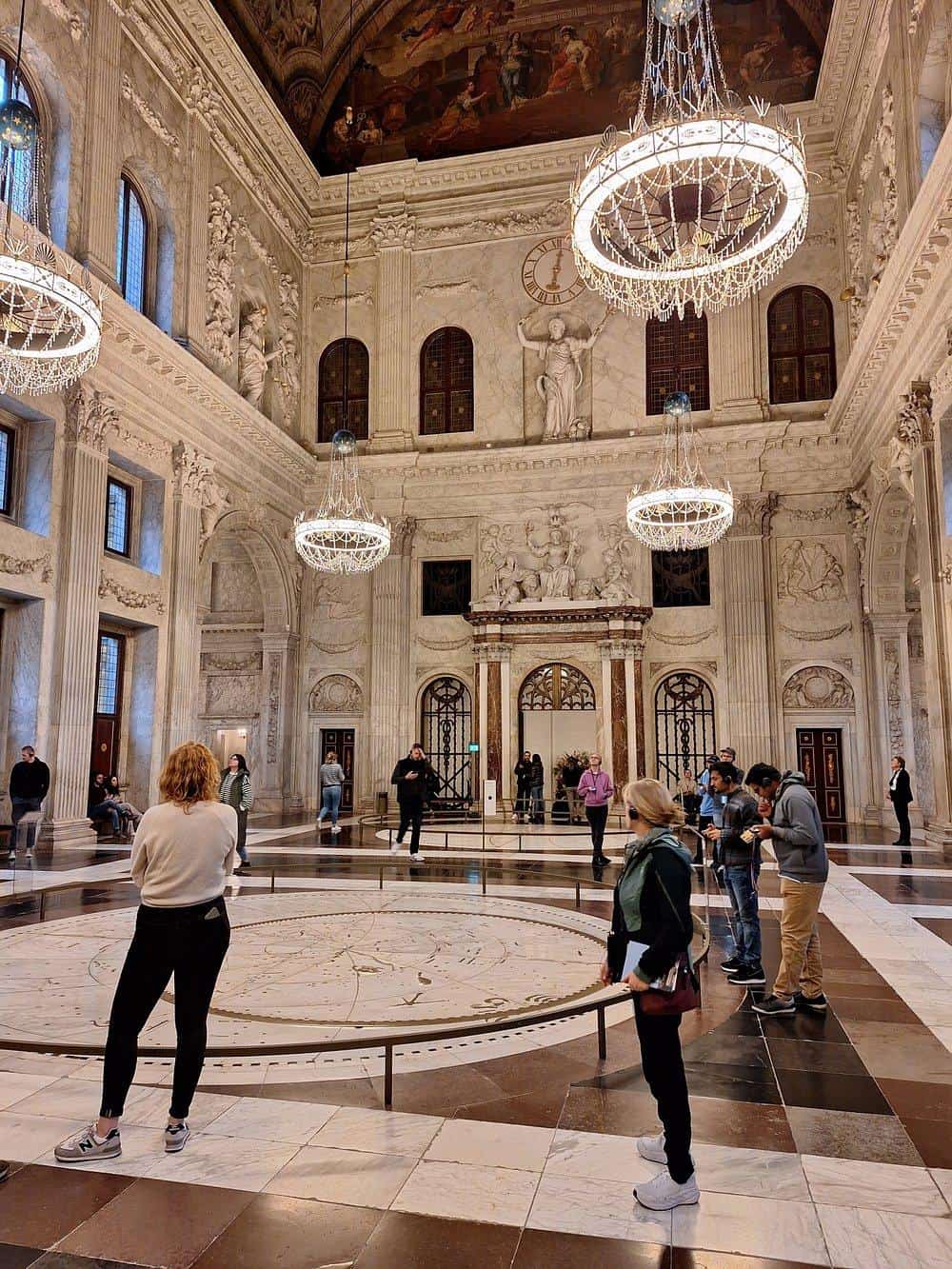 Elegant marble interior of historic museum with chandeliers and visitors exploring exhibits.