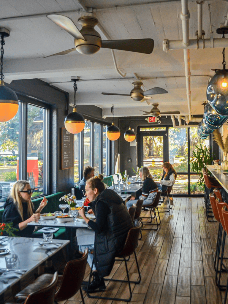 Cozy restaurant interior with large windows, stylish lighting, and diners enjoying meals, creating inviting dining ambiance.