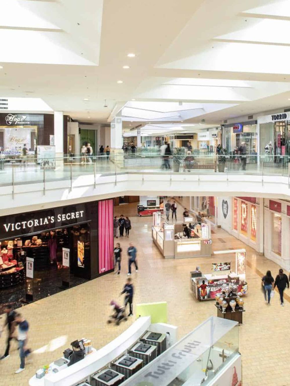 Bright shopping mall interior with stores, shoppers, and a Victoria’s Secret storefront.