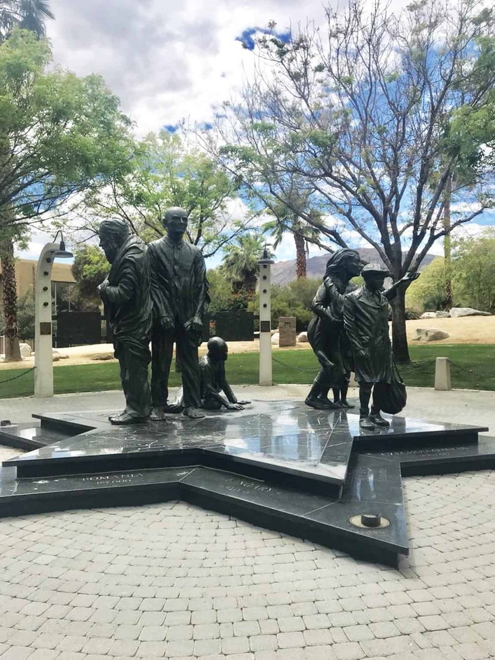 Bronze statue commemorating diverse individuals with desert landscaping and palm trees in background.