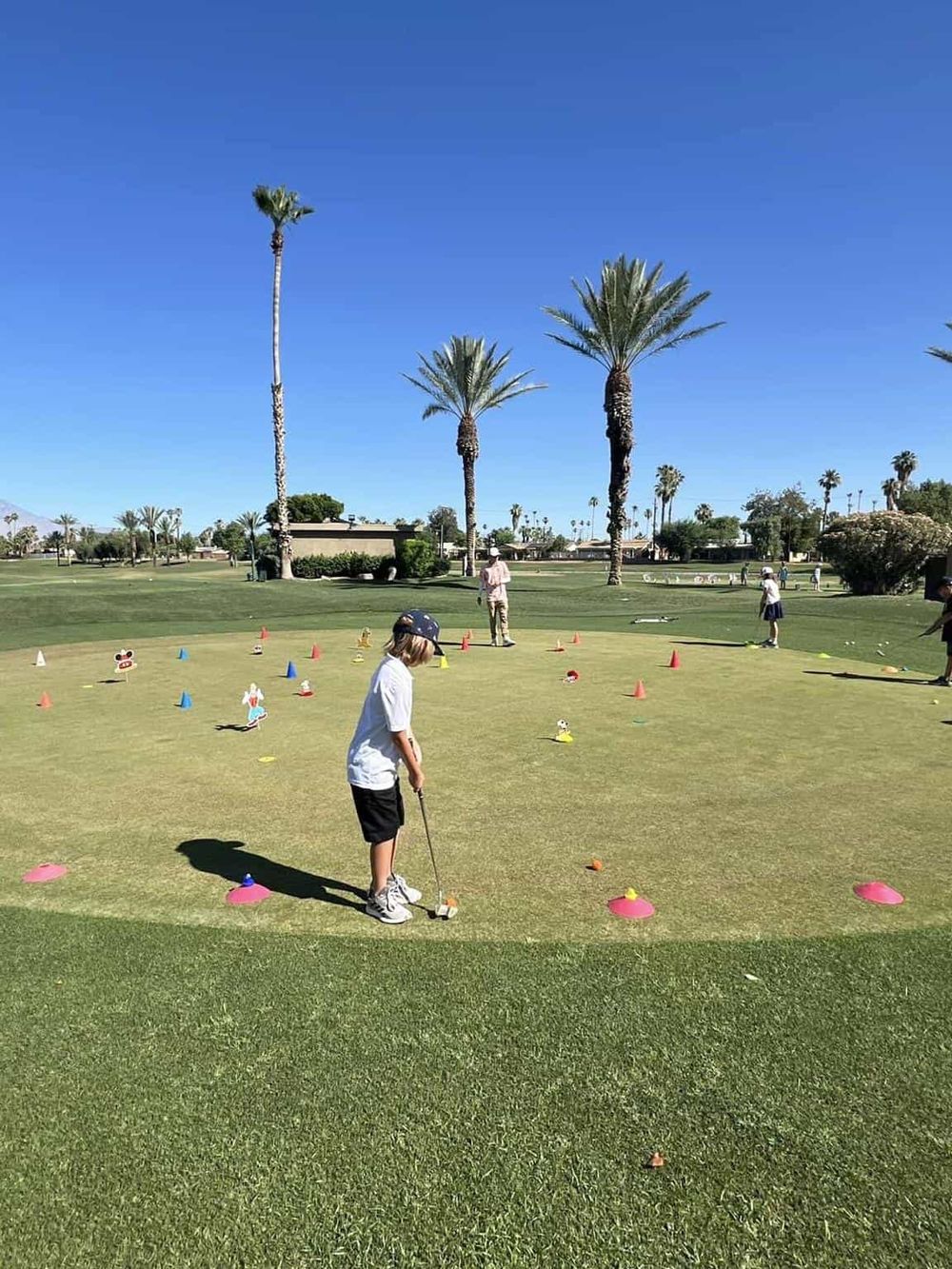 Child practicing golf on a driving range with palm trees and a clear blue sky in the background.