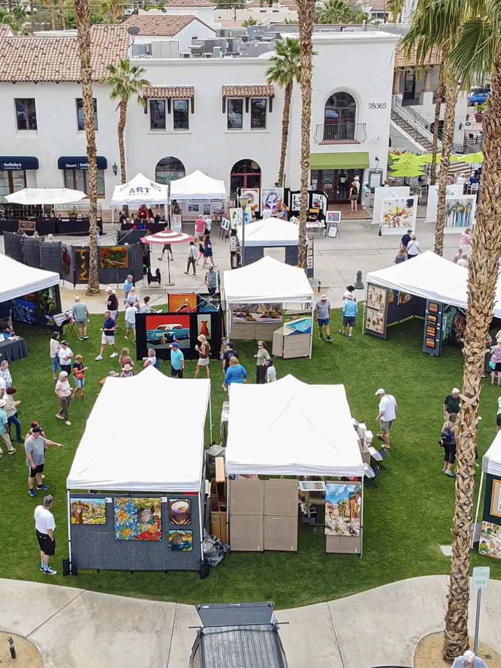 Colorful outdoor art market with tents, paintings, and visitors, set against a sunny urban backdrop.