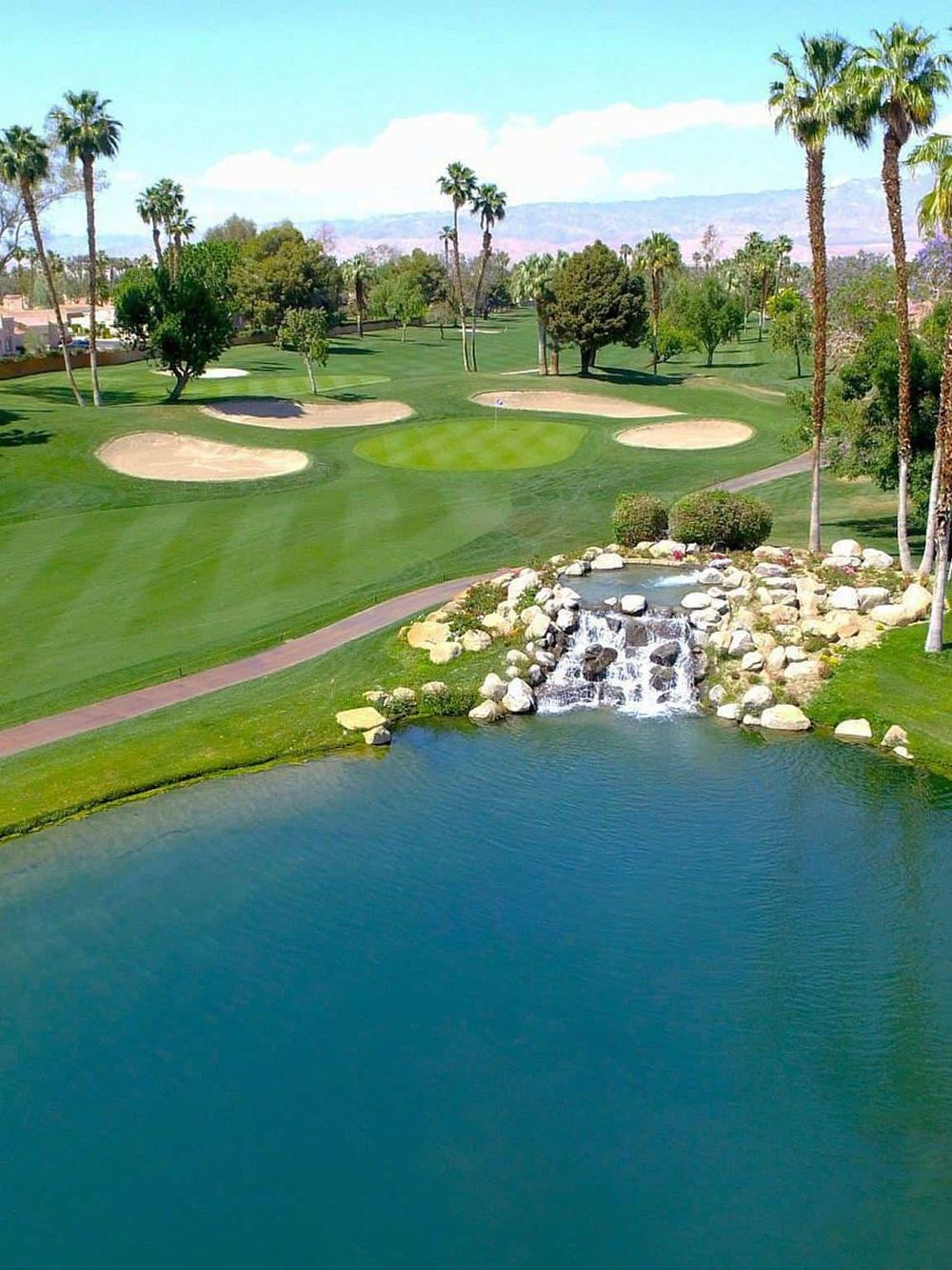 Lush golf course with water features, palm trees, and scenic mountain views during daytime.