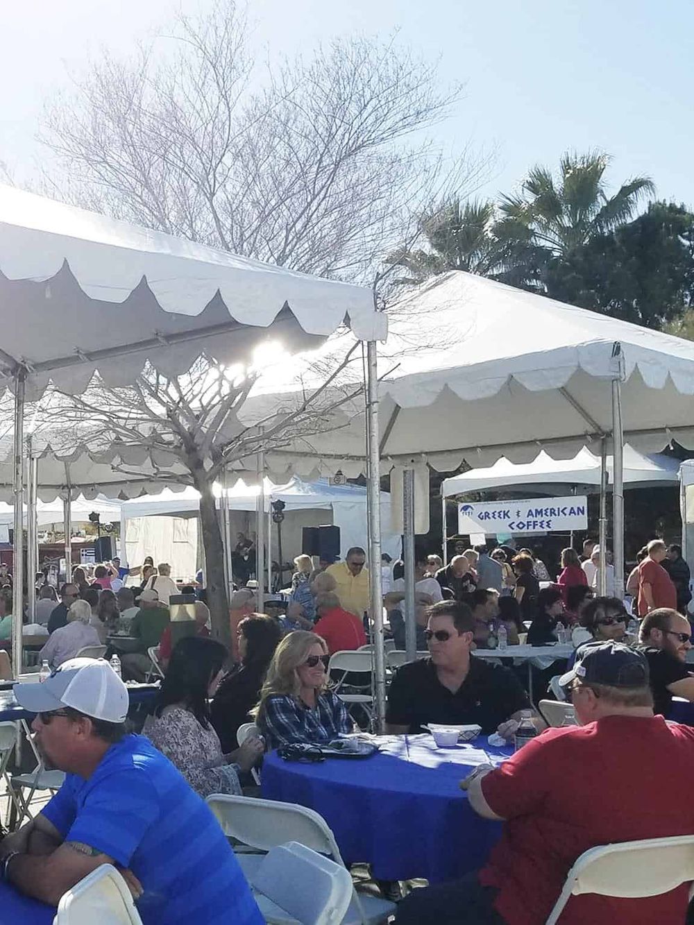 Vibrant outdoor event with diverse crowd, Greek and American coffee booths, and festive tents, showcasing community gathering and cultural celebration.