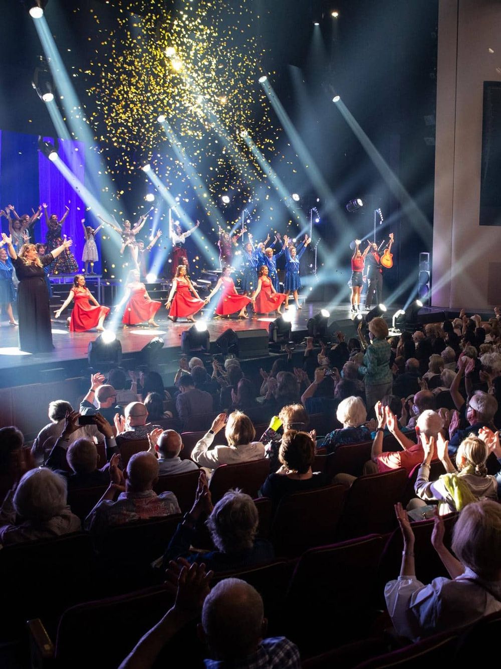 Bright stage performance with dancers, colorful costumes, and concert lighting in a theater setting.