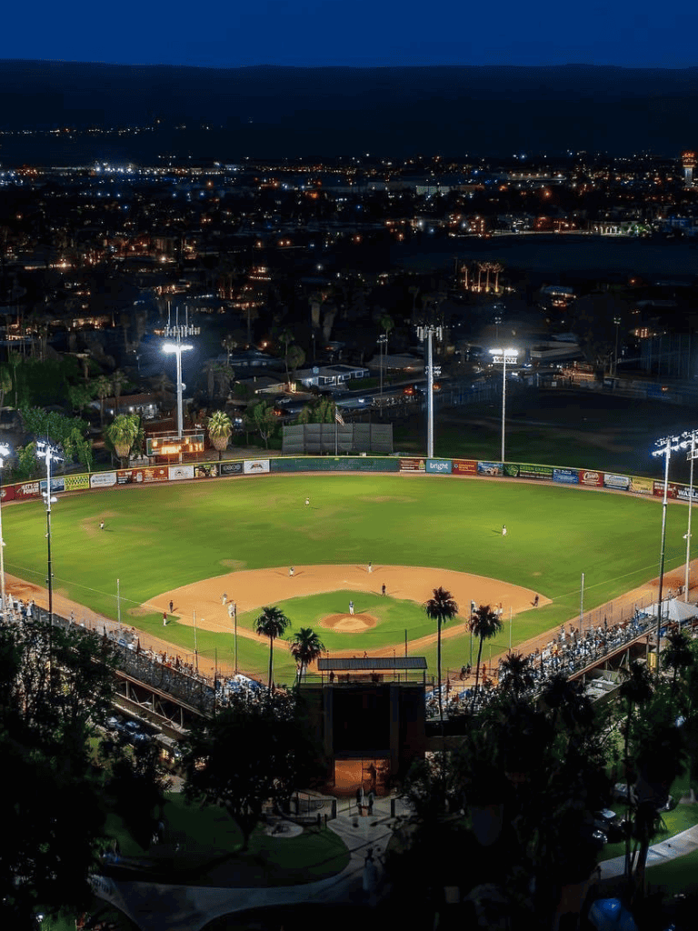 Nighttime view of a baseball stadium illuminated with bright lights, overlooking a cityscape with buildings and water body in the background.