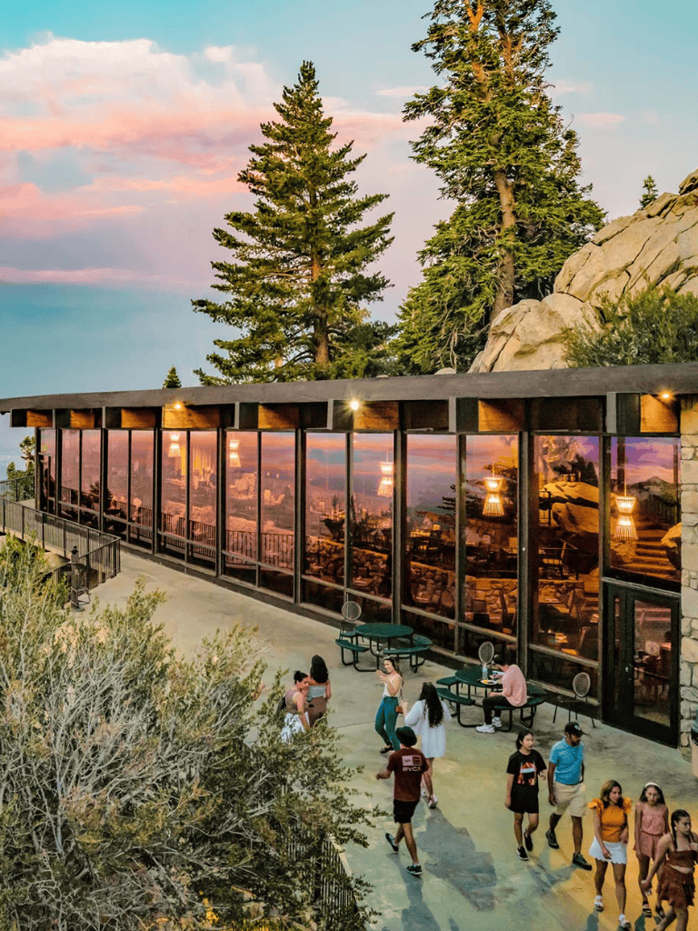 Spectacular mountain view with a modern glass building, lush trees, and visitors enjoying the scenic outdoor patio.