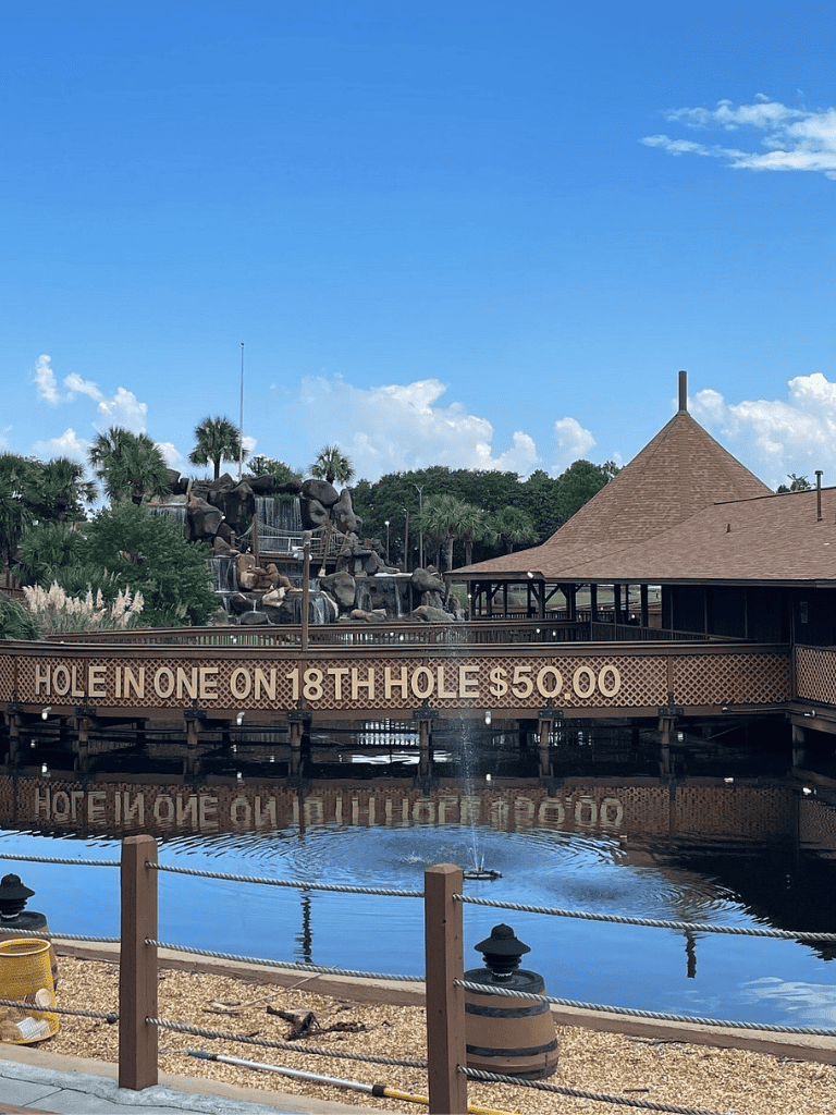 Shark-themed mini golf course near water with palm trees, blue sky, and a sign for hole 18.