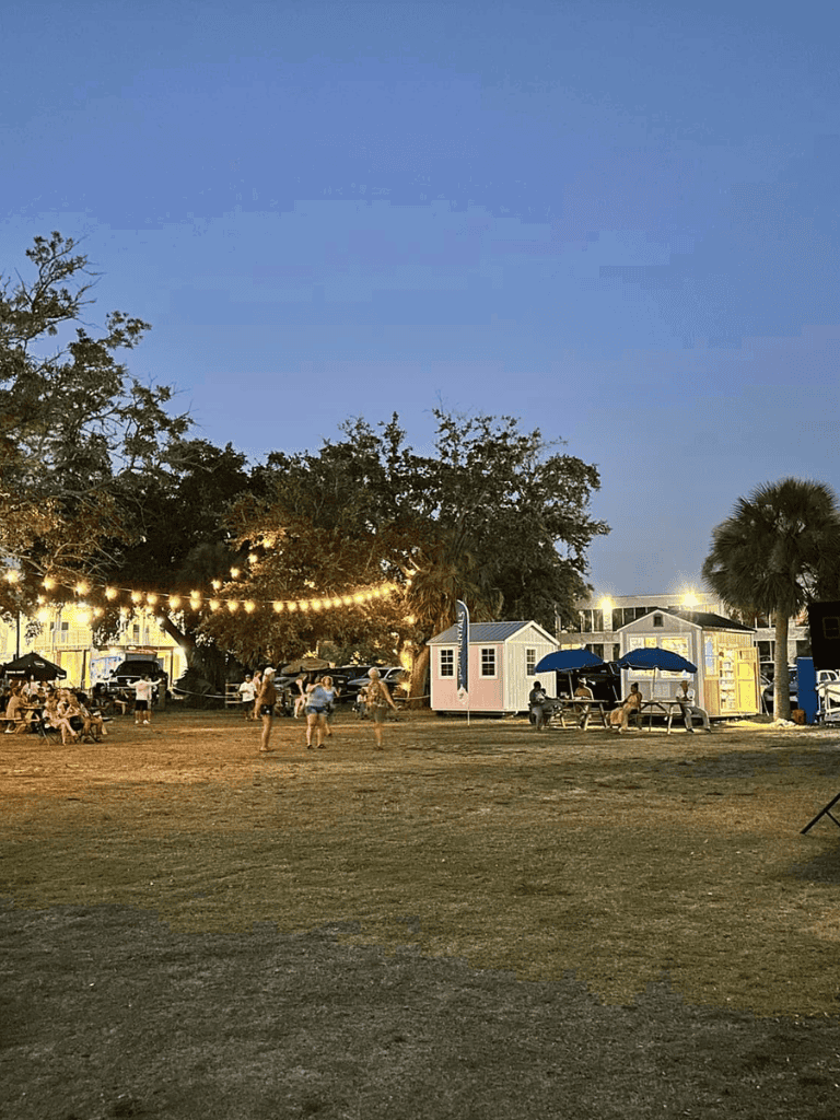 Booth area with string lights at dusk, outdoor event, food trucks, and seating, community gathering, family-friendly atmosphere.