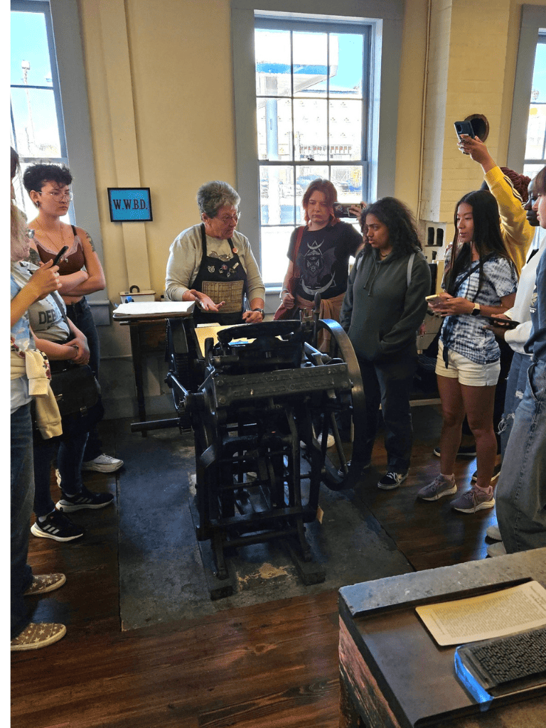 Museum tour guide explaining historic printing press to group of students, indoor setting.