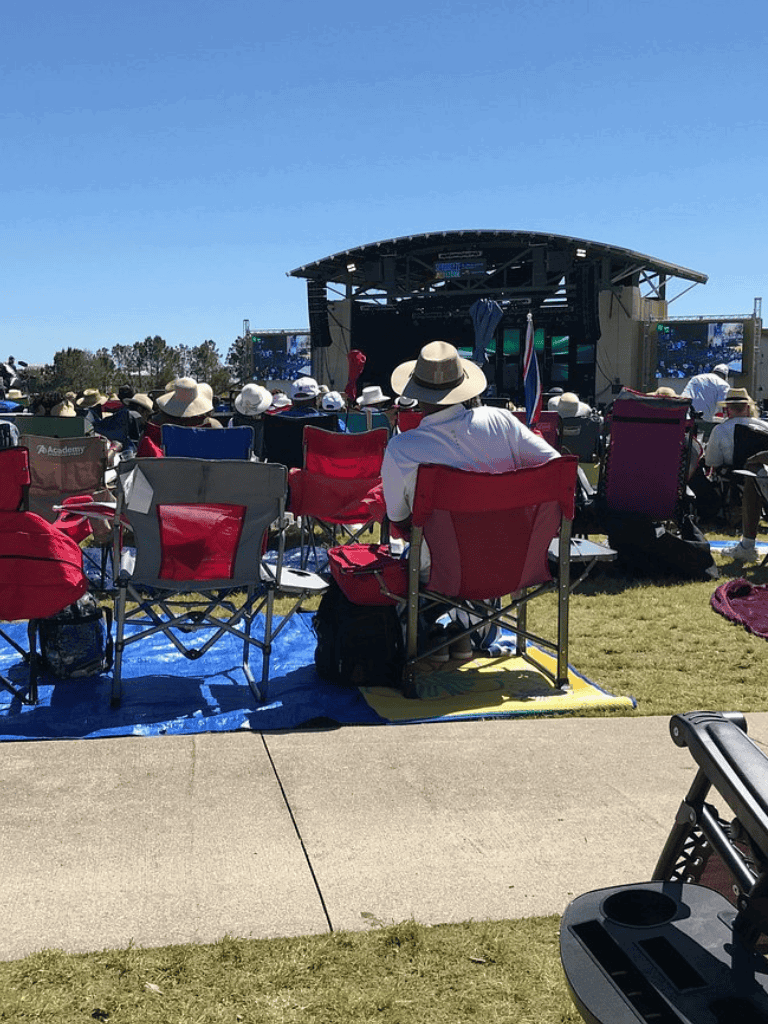 Crowd gathered at outdoor concert with stage, sitting on portable chairs, sunny day, enjoying live music.