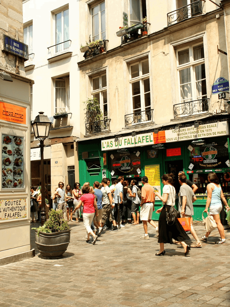 French street scene with people walking past a falafel shop, charming buildings, and a lively atmosphere.