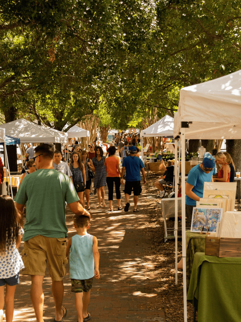 Vibrant outdoor market scene with diverse shoppers, vendors, and lush shaded trees, perfect for community and shopping events.