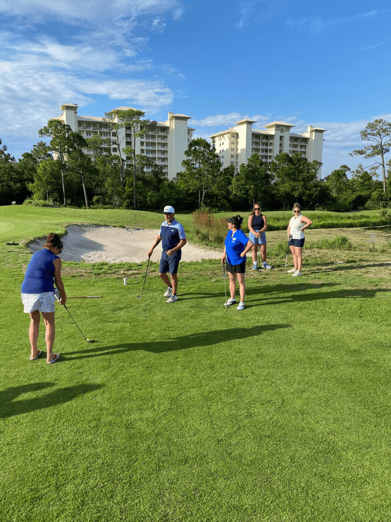 Woman playing golf on lush course with group of friends, scenic resort in background, sunny day.