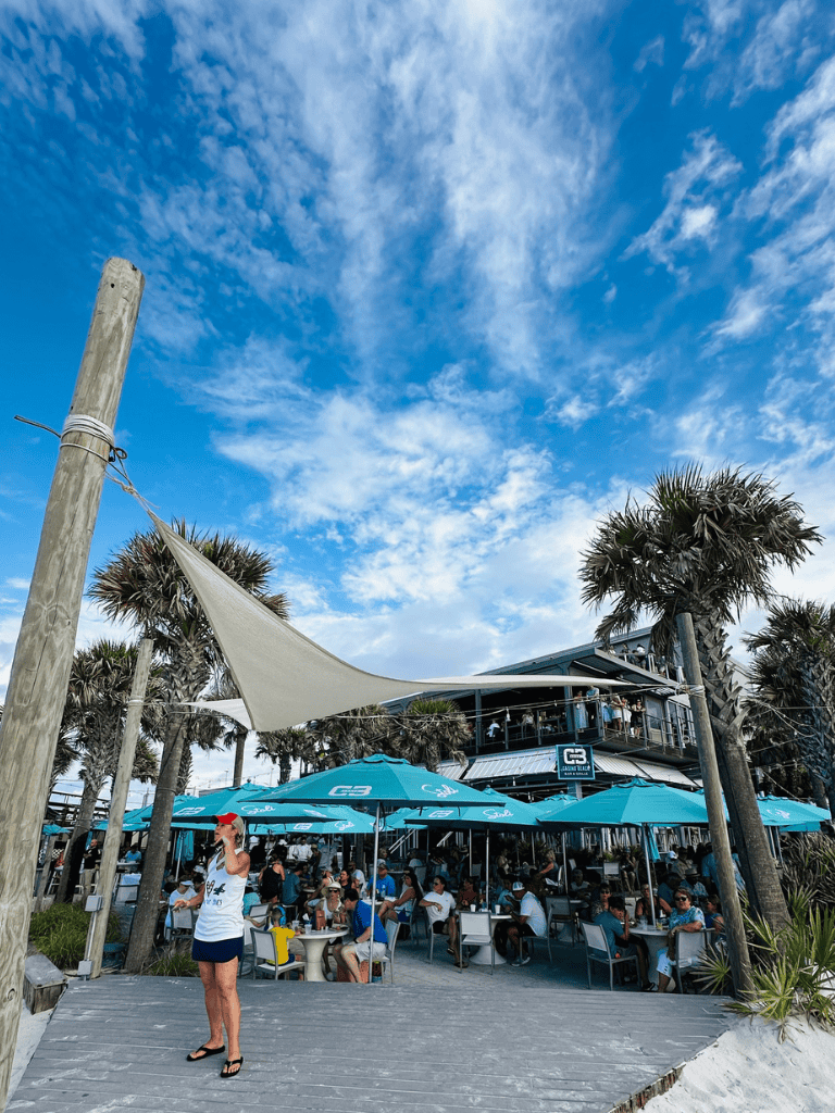 Relaxed outdoor beachside restaurant with crowd, umbrellas, and palm trees under blue sky.