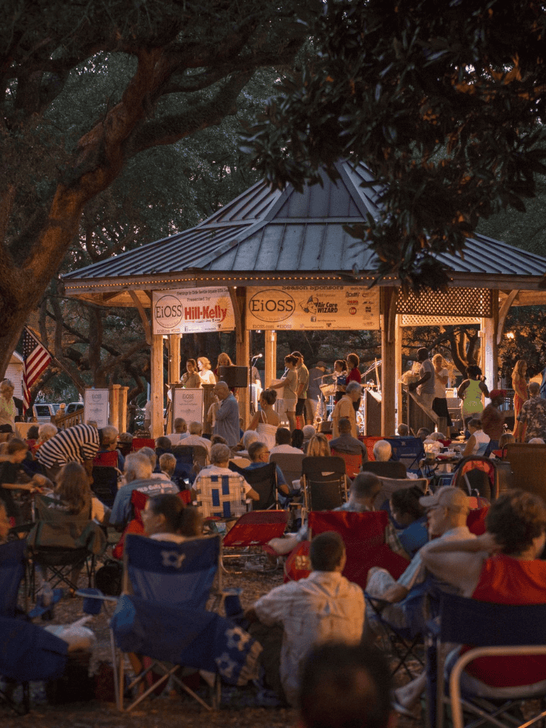 Music concert in a park at sunset, outdoor stage with audience, community event, family-friendly entertainment, summer evening celebration.
