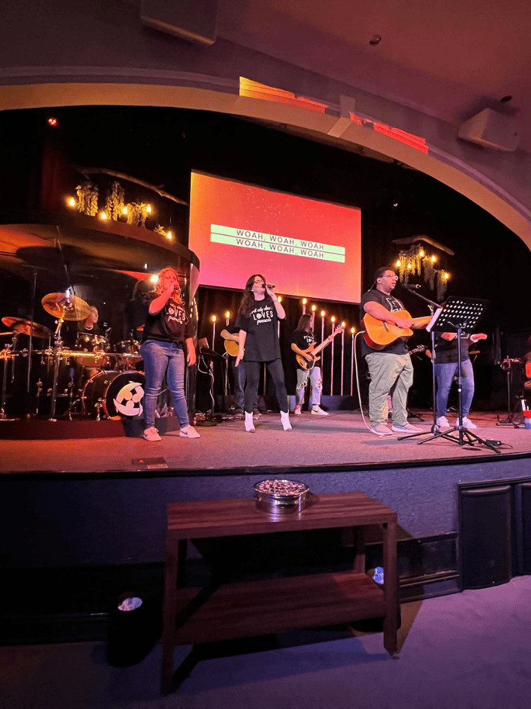 - Live band performing worship music with lyrics "WOAH, WOAH, WOAH" on large screen.